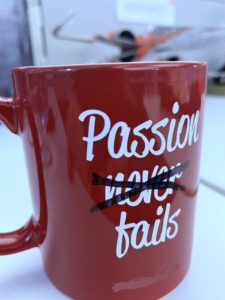 A red coffee mug with the words "Passion Never Fails" emblazoned on it has the word "Never" scratched out with felt pen.