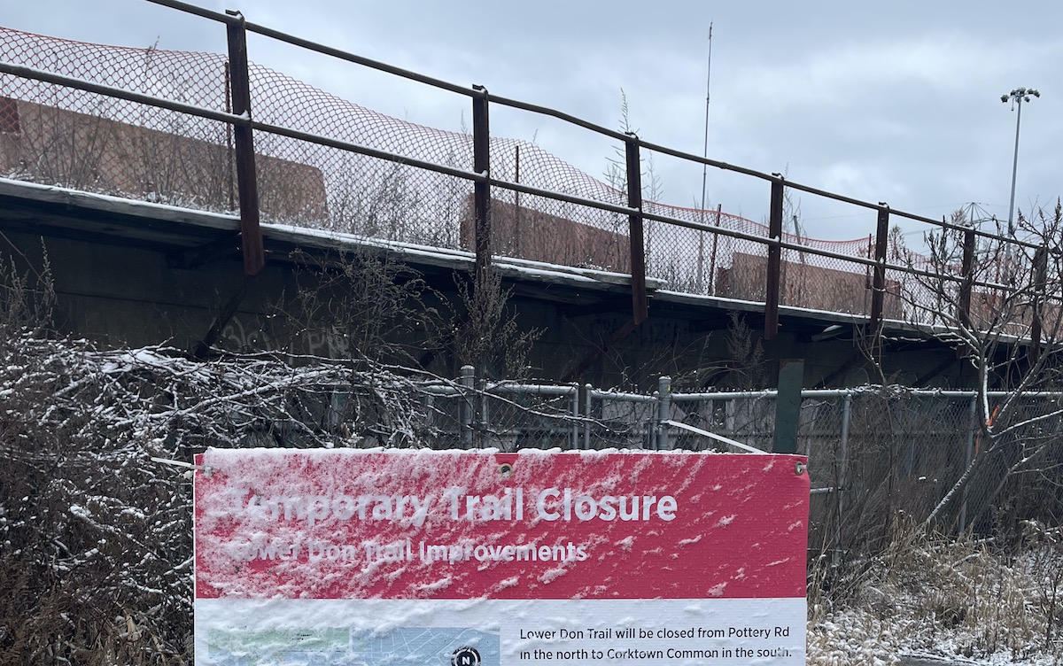 A public notice in Corktown, beneath the rail overpass, reading "Temporary Trail Closure." It is almost completely unreadable thanks to snow and graffiti.