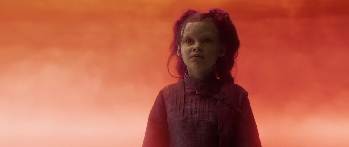 Young Gamora, a green-skinned alien girl, stands in an orange landscape, looking beseechingly at her father Thanos.