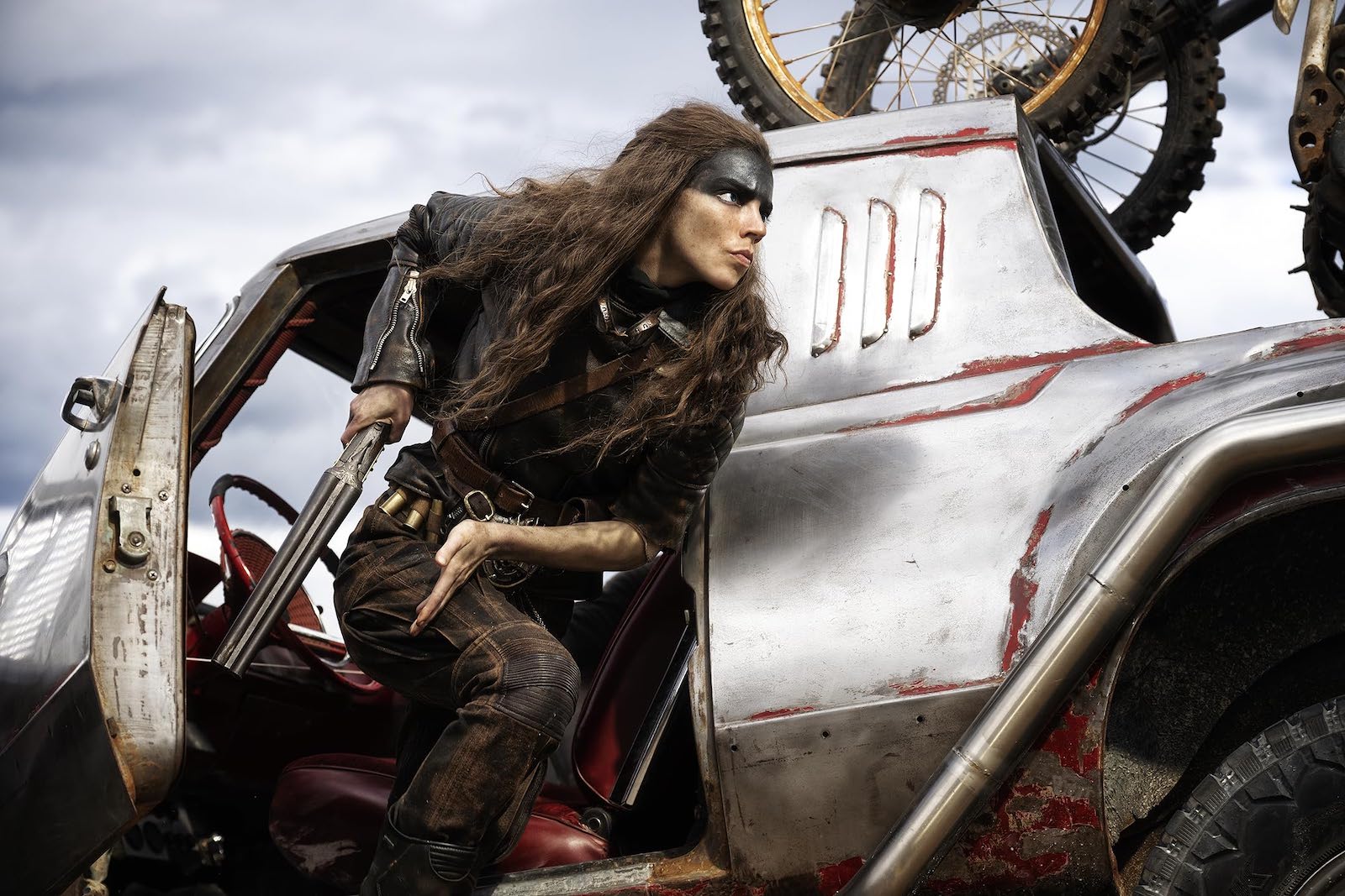 Furiosa, a leather-clad wasteland warrior with blue-black makeup covering her forehead, leaps from a car with gun in hand.