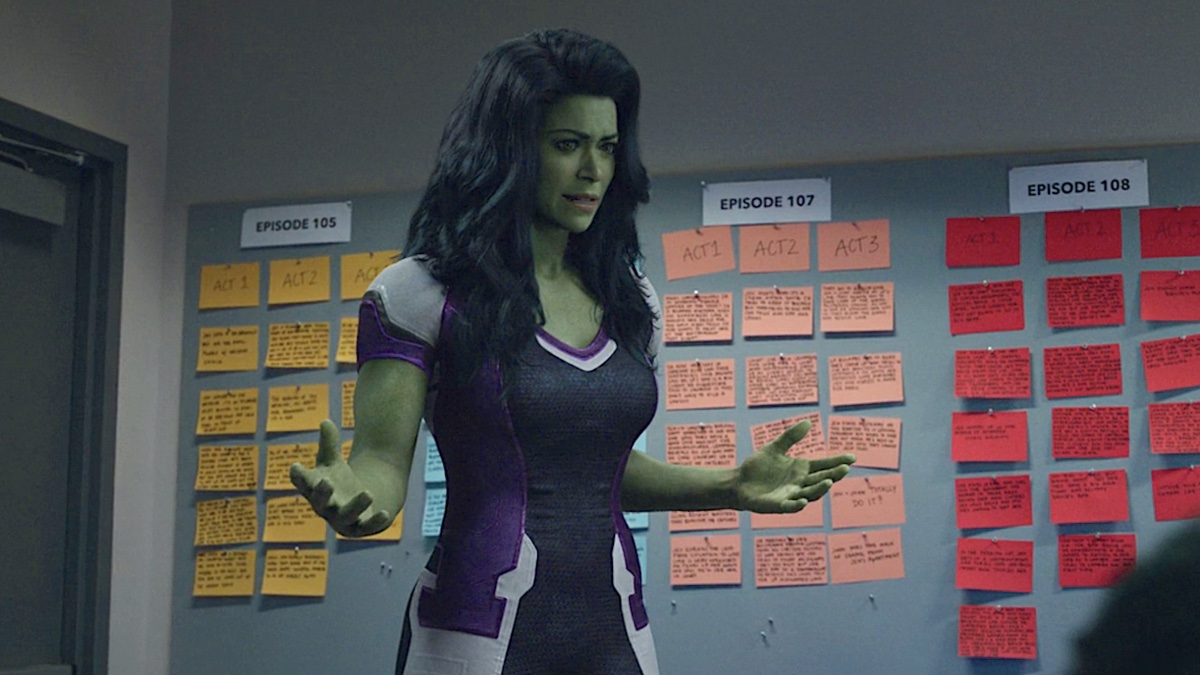 She-Hulk, an enormous green-skinned woman in a purple and white unitard, stands in front of a television writers' beat board.