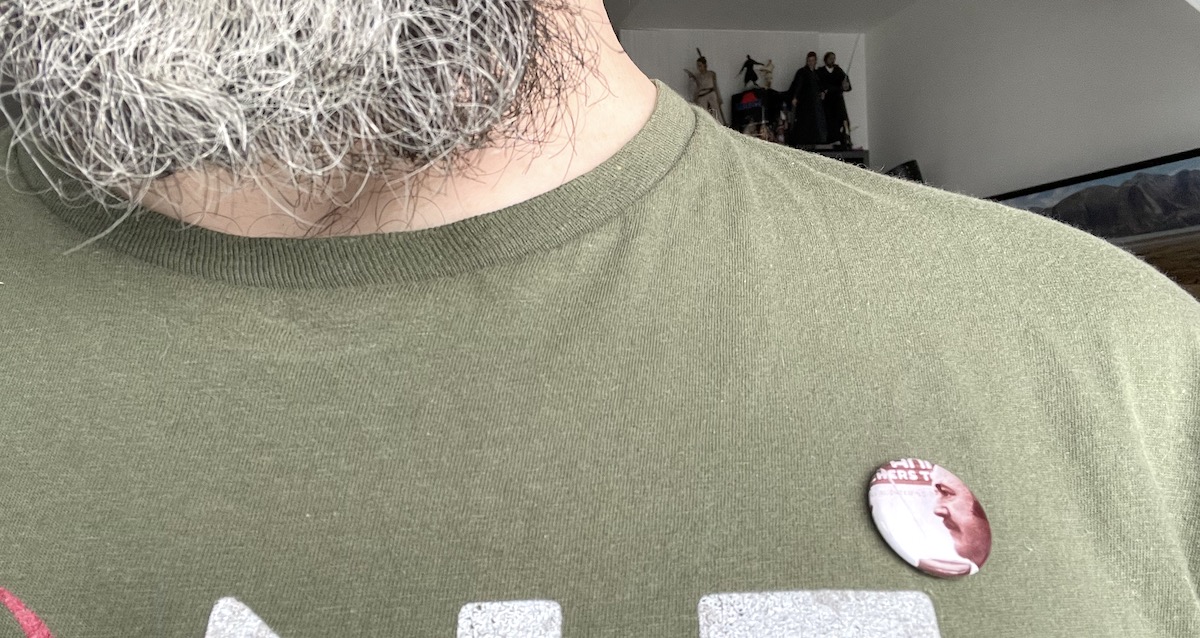 A homemade button of Gene Hackman, starring in the 1972 film "Prime Cut," adorns a green t-shirt on a grey-bearded man.